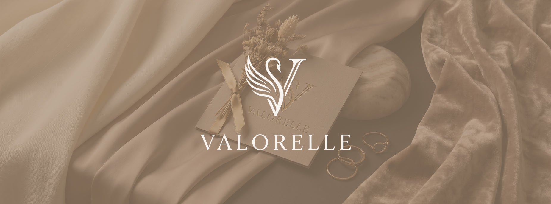 A beige and brown monochrome styled flat-lay featuring a square card with a white winged 'V' logo and the brand name 'VALORELLE' centered on the image, surrounded by draped satin fabric, dried flowers, and rings.